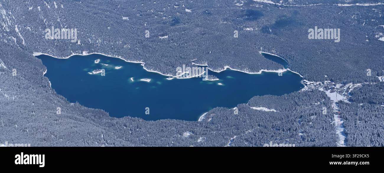 Un colpo aereo panoramico ad angolo alto del profondo lago blu dell'Eibsee con le sue piccole isole, completamente circondato da una fitta foresta di pini innevata in Foto Stock