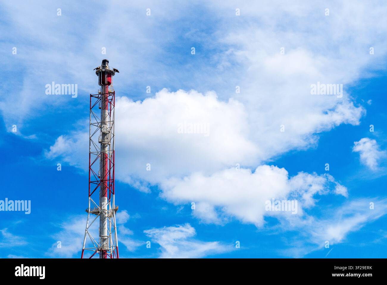Pila rossa e bianca di un impianto petrolchimico che si erge contro un cielo blu vivace con nuvole bianche, utilizzata per bruciare il gas in eccesso nell’industria petrolifera Foto Stock