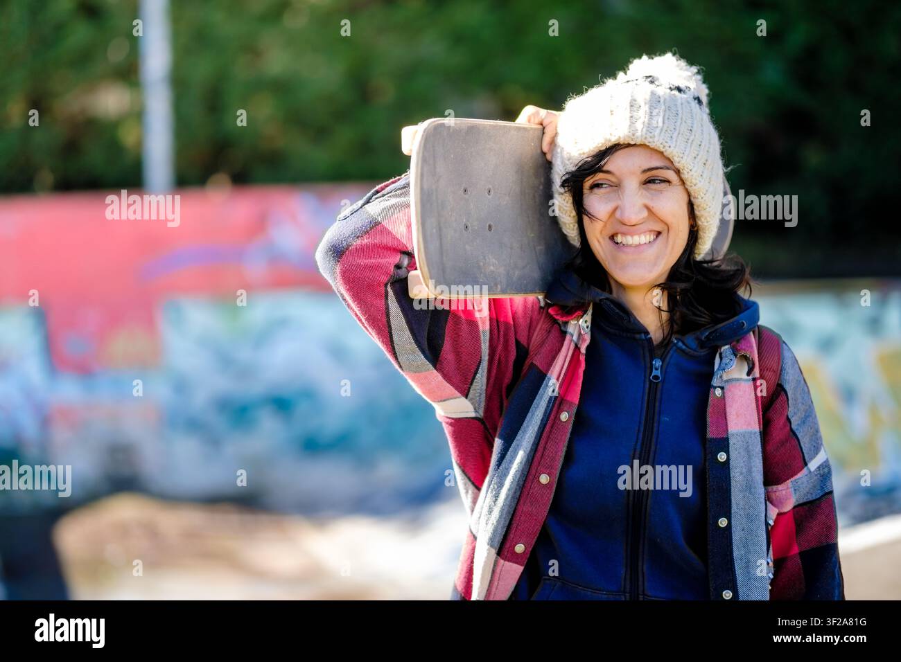Giovane donna sorridente che tiene uno skate sulla spalla in uno skatepark all'aperto Foto Stock