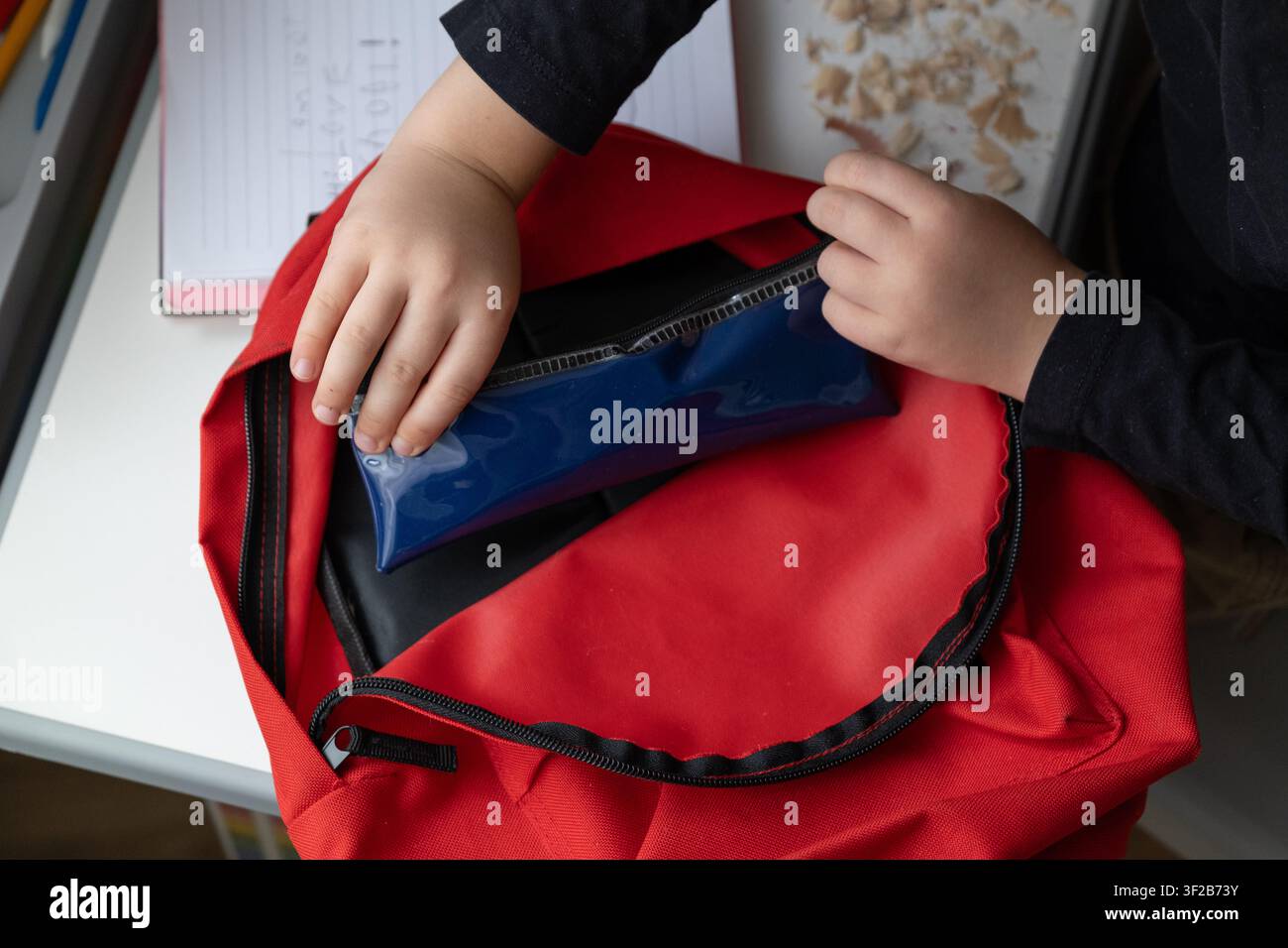 Vista dall'alto di un bambino che mette una trousse in uno zaino scolastico aperto mentre si prepara per la scuola. La scena realistica domestica illustra l'organizzazione sch Foto Stock