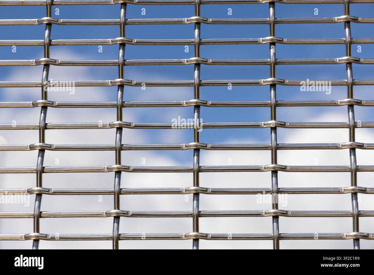 Primo piano di rete metallica in acciaio inossidabile tessuta con cielo blu e nuvole morbide visibili attraverso le aperture. Foto Stock