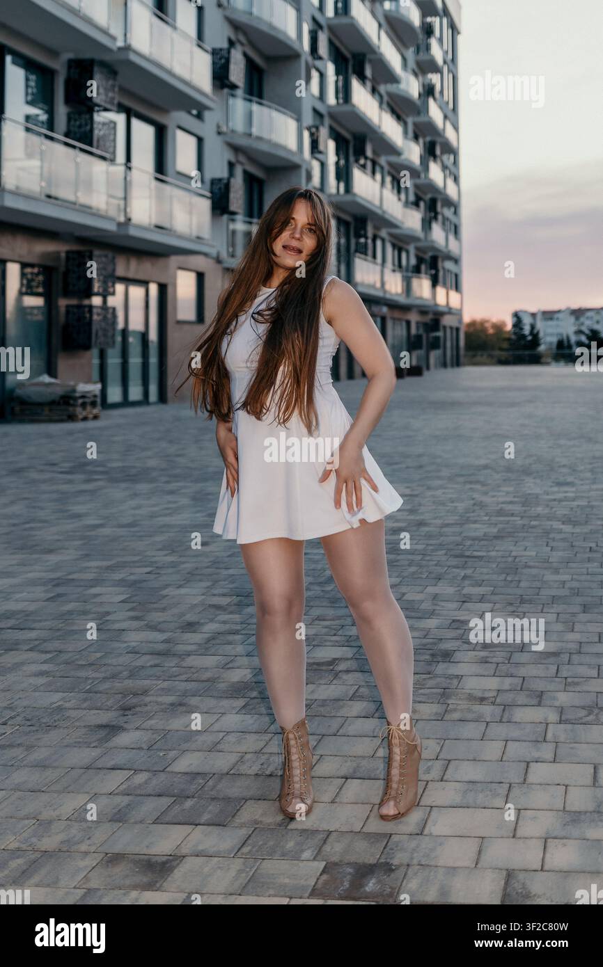Donna fashion city in posa con vestito bianco vicino a edifici residenziali moderni durante la luce del tramonto serale Foto Stock