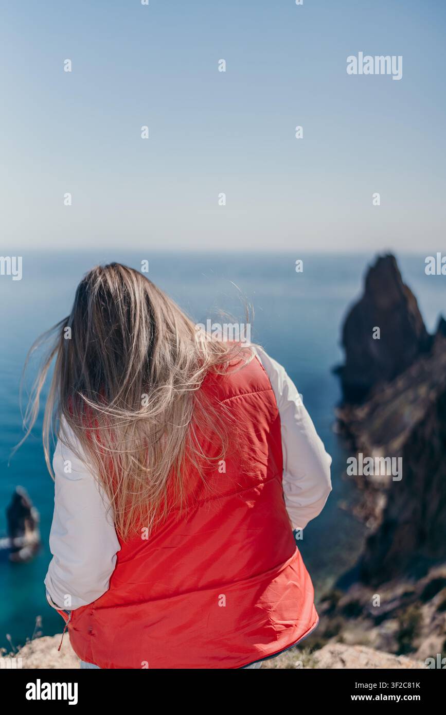 Donna su una scogliera sul mare Una donna in un giubbotto rosso si trova sul bordo di una montagna rocciosa che si affaccia sul vasto oceano blu in una giornata di sole Foto Stock