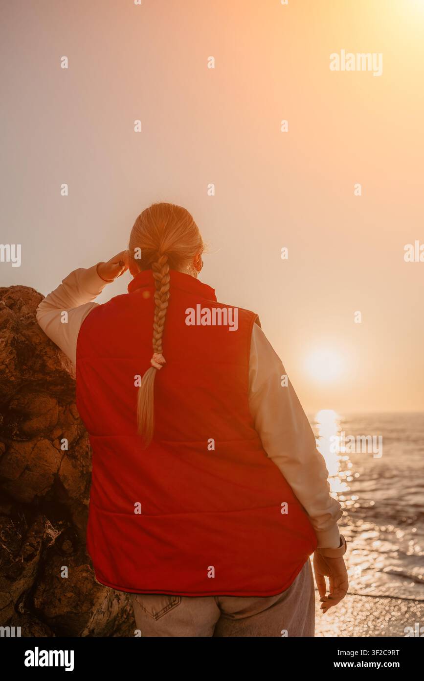 Donna al tramonto sull'oceano si trova sulla costa indossando un giubbotto rosso e guardando l'orizzonte durante l'ora dorata sulla spiaggia Foto Stock