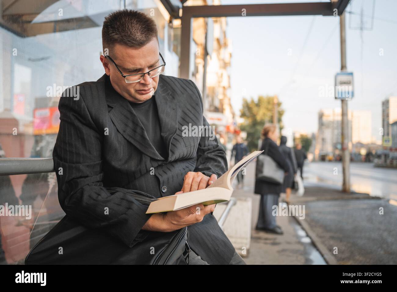 Uomo con gli occhiali che legge un libro aperto mentre è seduto e aspetta l'autobus a una fermata dell'autobus urbana. Foto Stock