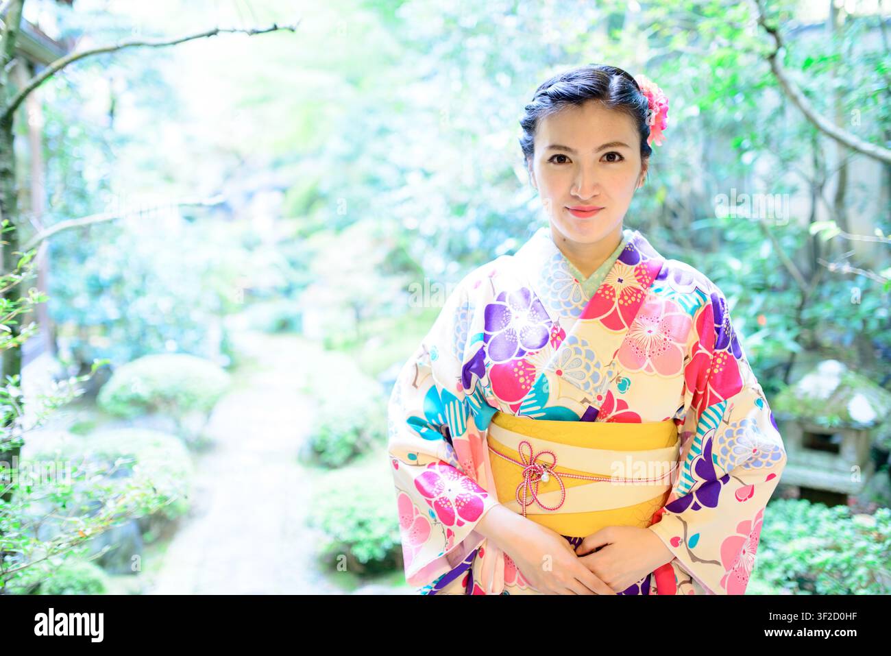 Una persona che indossa un kimono floreale colorato e un obi giallo si trova su un sentiero del giardino sfocato in Giappone. Foto Stock