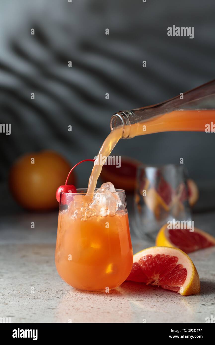Il succo di pompelmo viene versato in un bicchiere con ghiaccio. Bere in un bicchiere congelato e frutta fresca su un tavolo di pietra. Sullo sfondo, le ombre del palmo leav Foto Stock