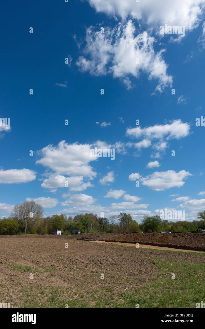 Ampio cielo primaverile blu con nuvole bianche cumulus sparse sopra un campo rurale appena arato e alberi lontani Foto Stock