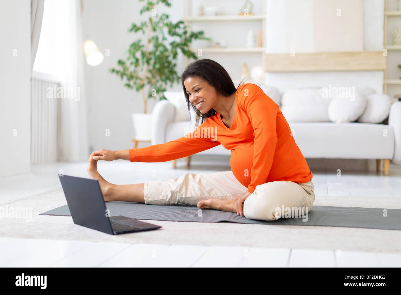 Donna nera incinta che si allena online, stirando la gamba su un tappeto da yoga a casa Foto Stock