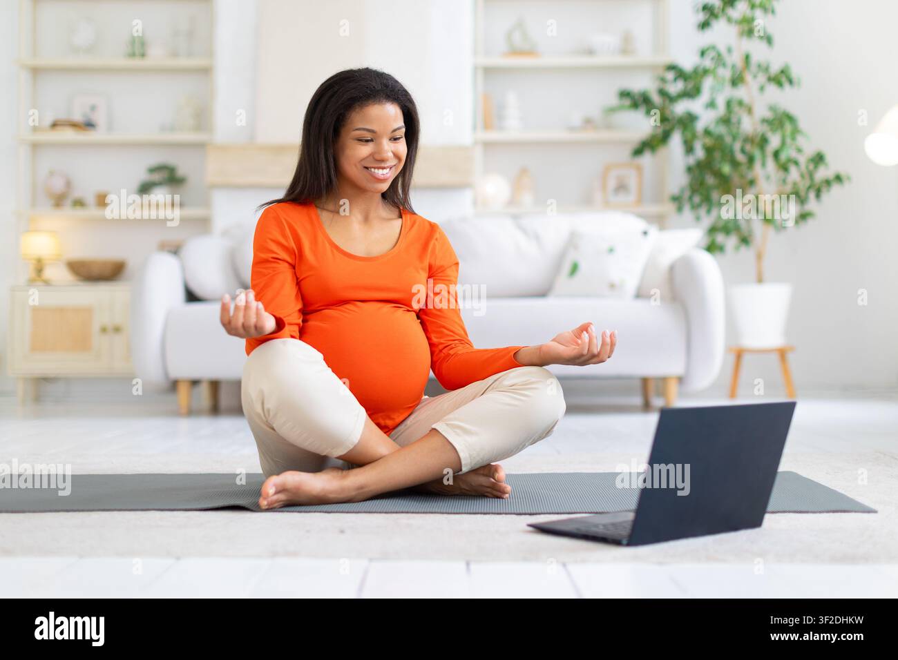 Donna nera incinta sorridente segue una sessione di meditazione online su laptop Foto Stock