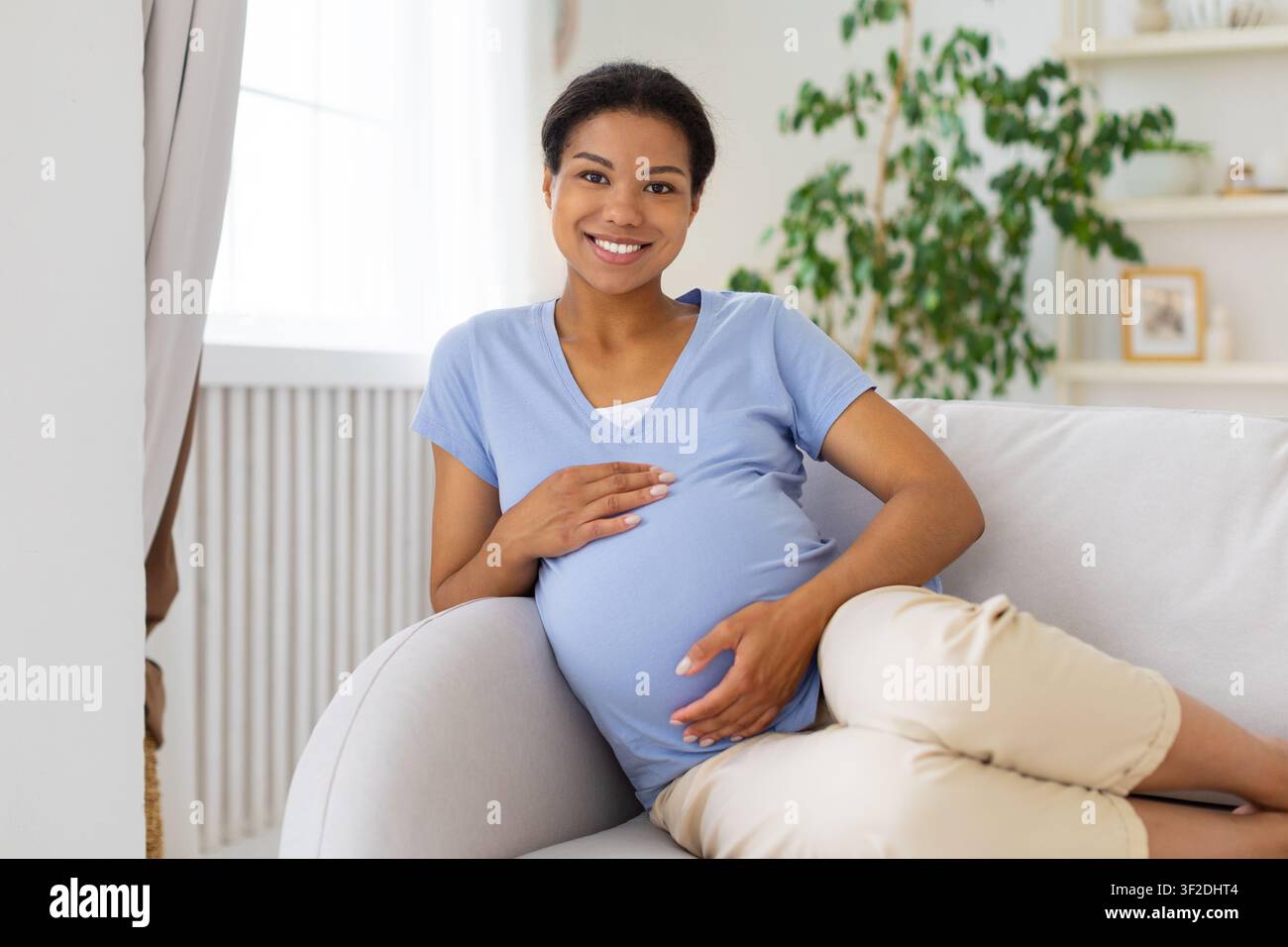 Donna nera incinta felice che si rilassa sul divano e tiene il ventre Foto Stock