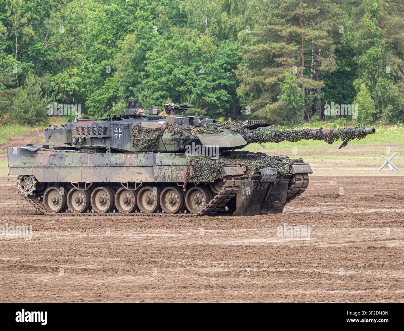Un carro armato principale tedesco Leopard 2 mimetizzato con l'equipaggio visibile nei portelli attraversa un campo di terra con uno sfondo di pineta. Foto Stock