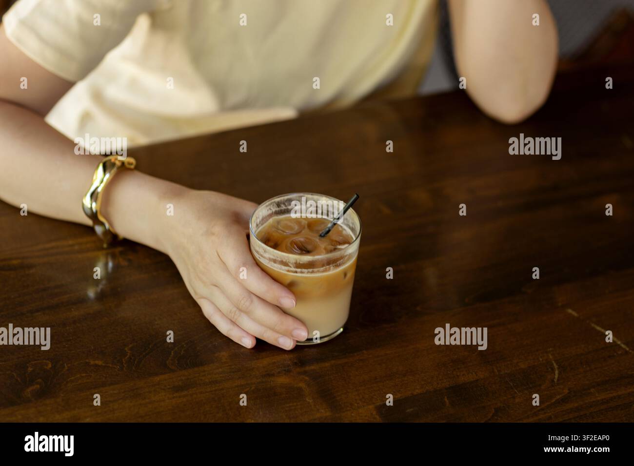 Una donna con un braccialetto d'oro tiene un caffè freddo in un caffè. Indossa una maglietta color crema, appoggiando il gomito sul tavolo di legno, creando un'atmosfera rilassata Foto Stock