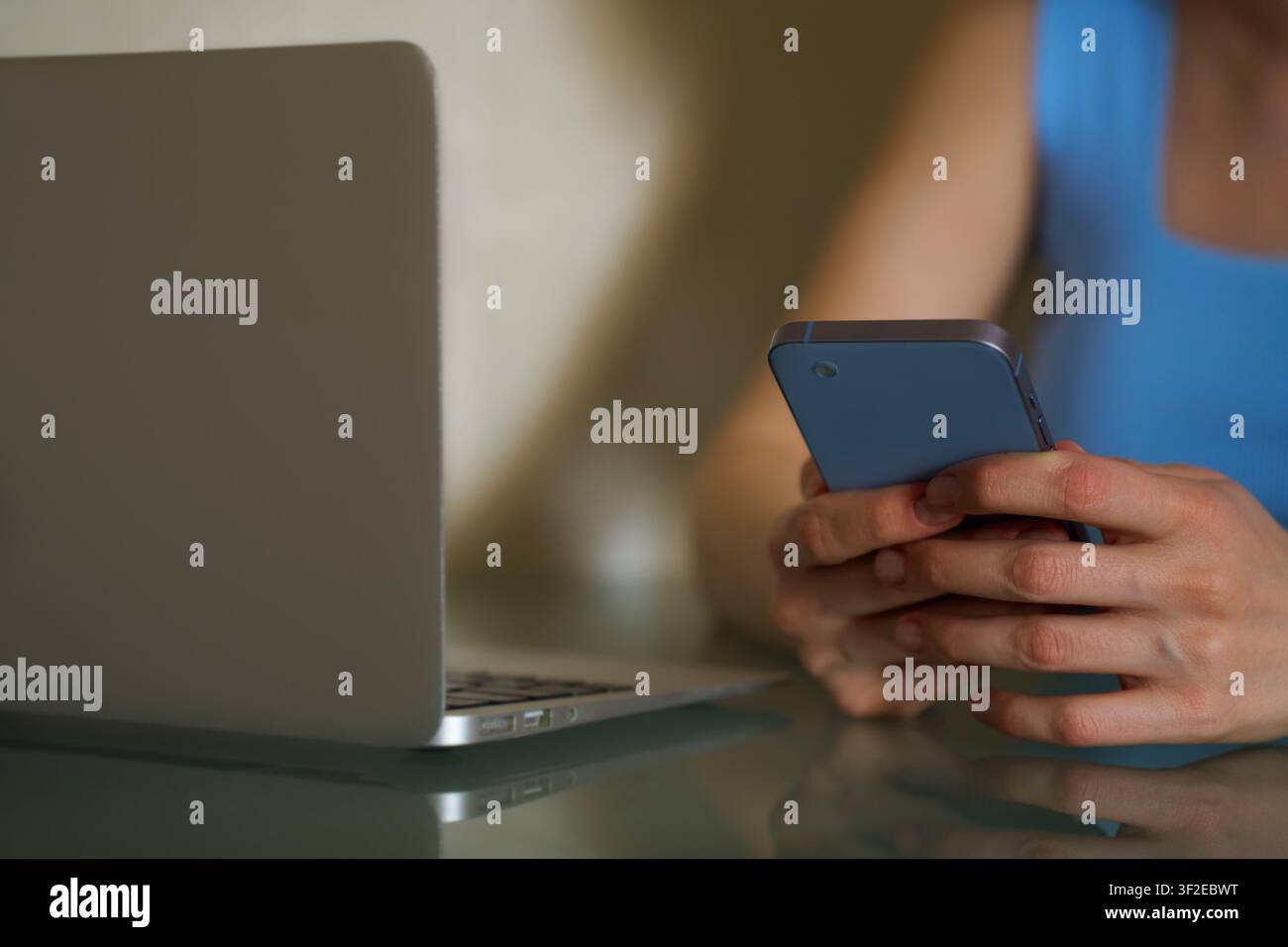 Una donna con una maglietta blu è immersa nel suo smartphone mentre un laptop è aperto accanto a lei. Questa immagine rappresenta il multitasking, la comunicazione online e Foto Stock