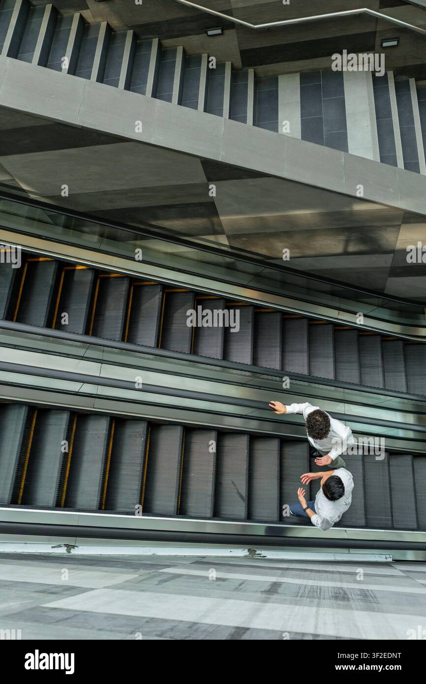 Vista dall'alto di due persone che percorrono scale mobili parallele all'interno di un moderno edificio in cemento e vetro, creando linee diagonali forti e una trama urbana astratta Foto Stock