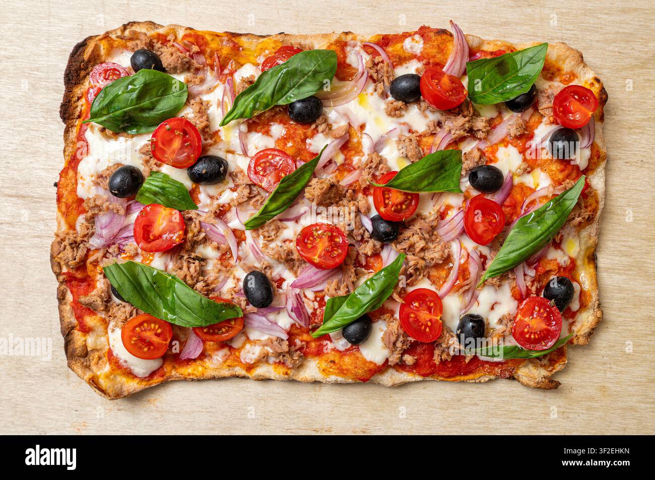Pinsa romana italiana guarnita con tonno, cipolla rossa, olive nere, pomodorini, mozzarella e basilico fresco, presentata su una tavola di legno Foto Stock