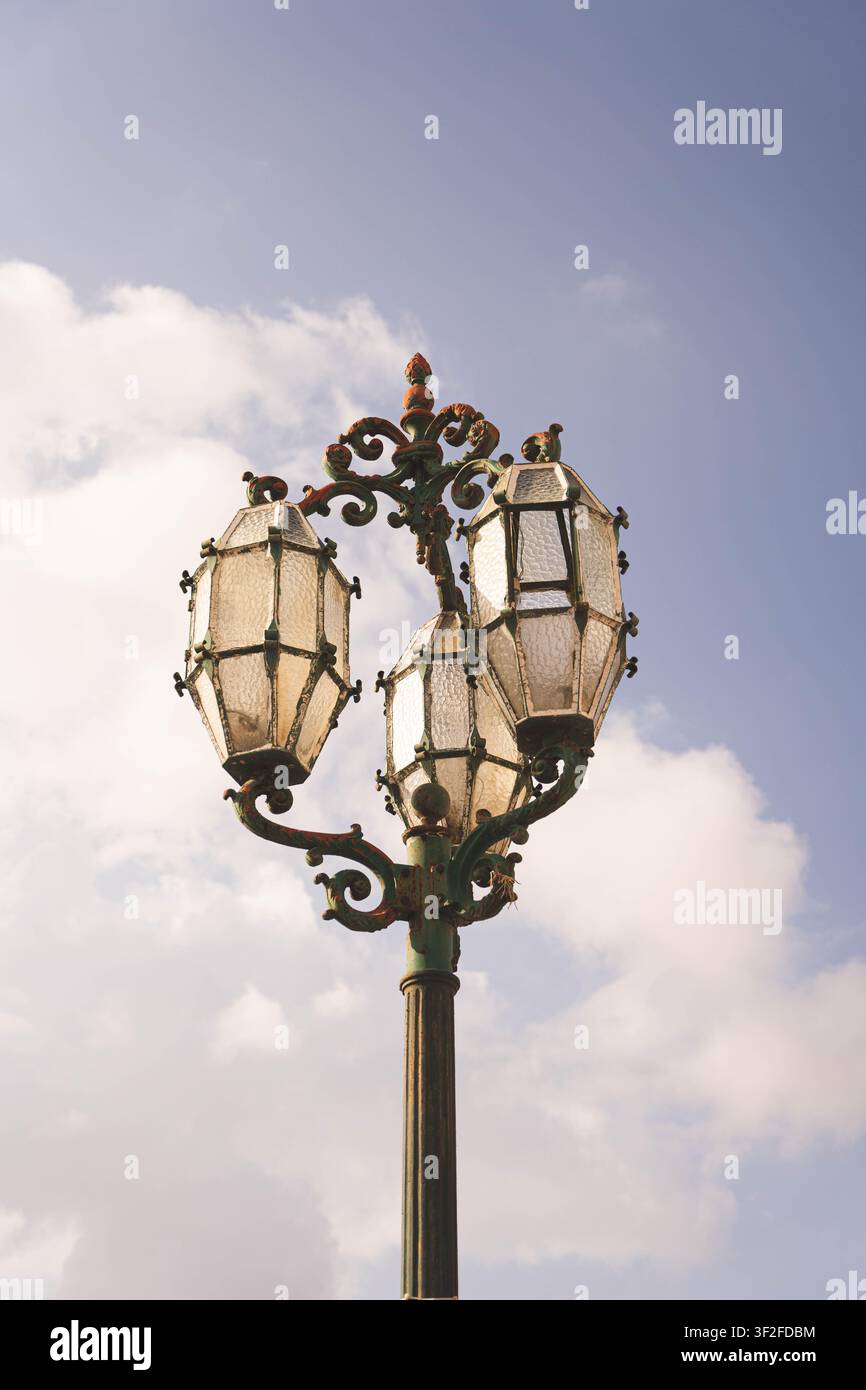Valletta, Regione del Porto, Malta – 14 luglio 2022: Vista di un lampione vintage ornato con tre lanterne in vetro contro un cielo blu con nuvole morbide. Foto Stock