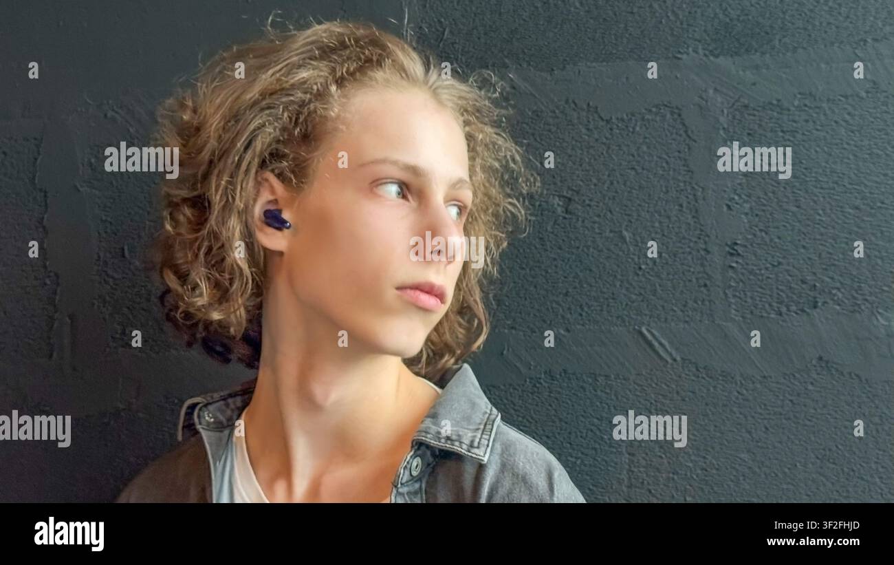 Adolescente pensieroso con auricolari senza fili che guarda lontano su uno sfondo minimalista scuro Foto Stock
