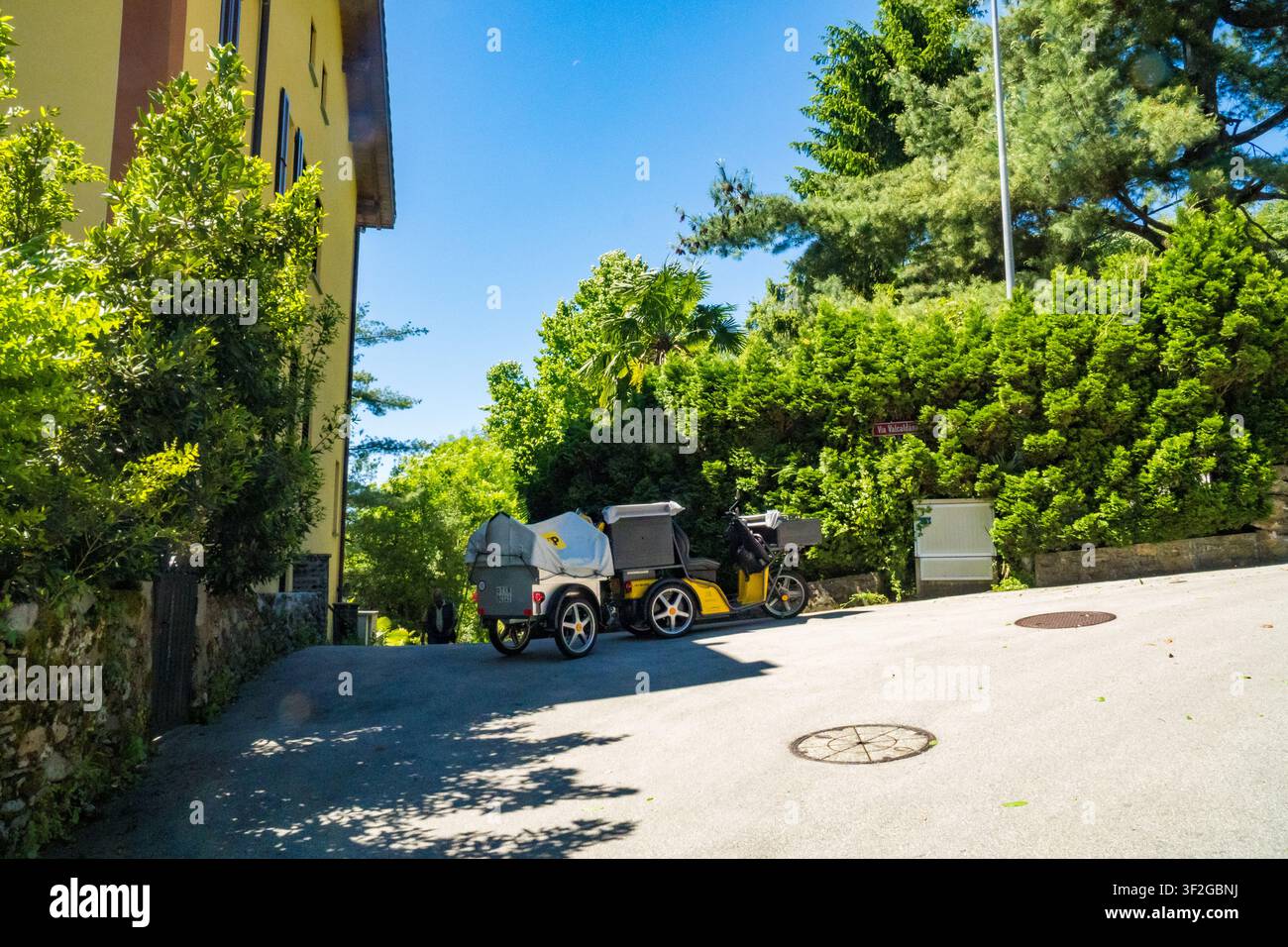Caslano - Svizzera - 12 maggio 2026: carrello di consegna della posta parcheggiato in una tranquilla strada di villaggio fiancheggiata da vegetazione e case, Malcantone, Ticino. Foto Stock