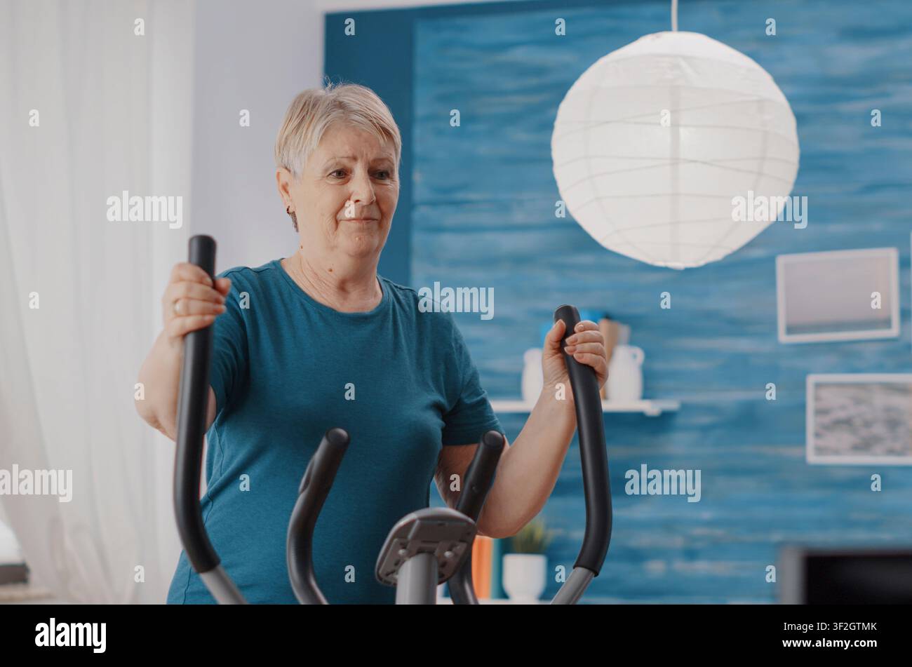 Donna in pensione pedala su una bicicletta stazionaria a casa, utilizzando una macchina cardio compatta per allenarsi, fare ciclismo e mantenere l'attività di fitness per anziani. Foto Stock