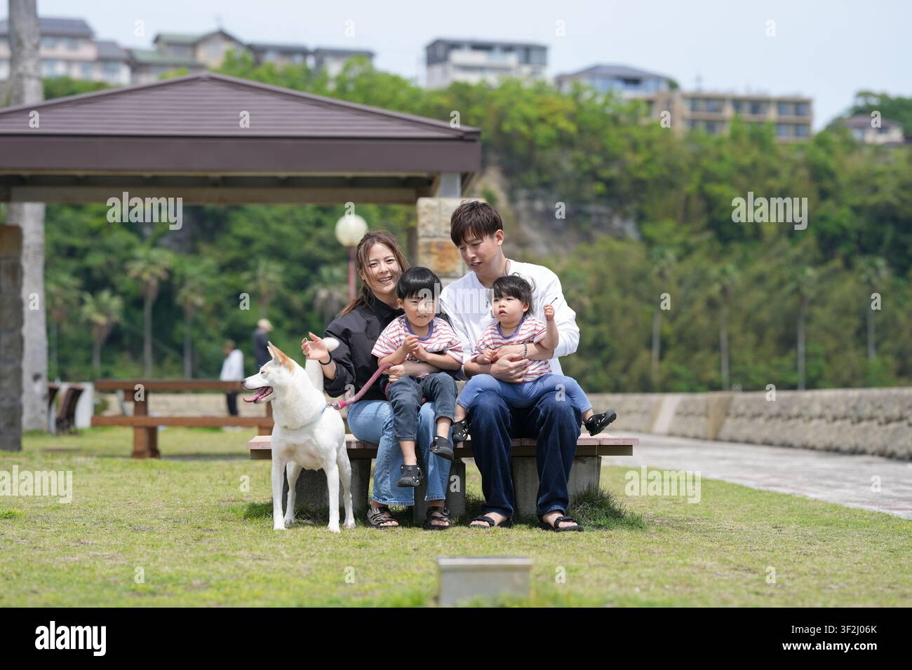 Famiglia seduta insieme su una panchina del parco con un cane bianco. La madre e il padre tengono stretti due bambini. Il cane è nelle vicinanze e guarda verso l'obiettivo. Erba verde Foto Stock