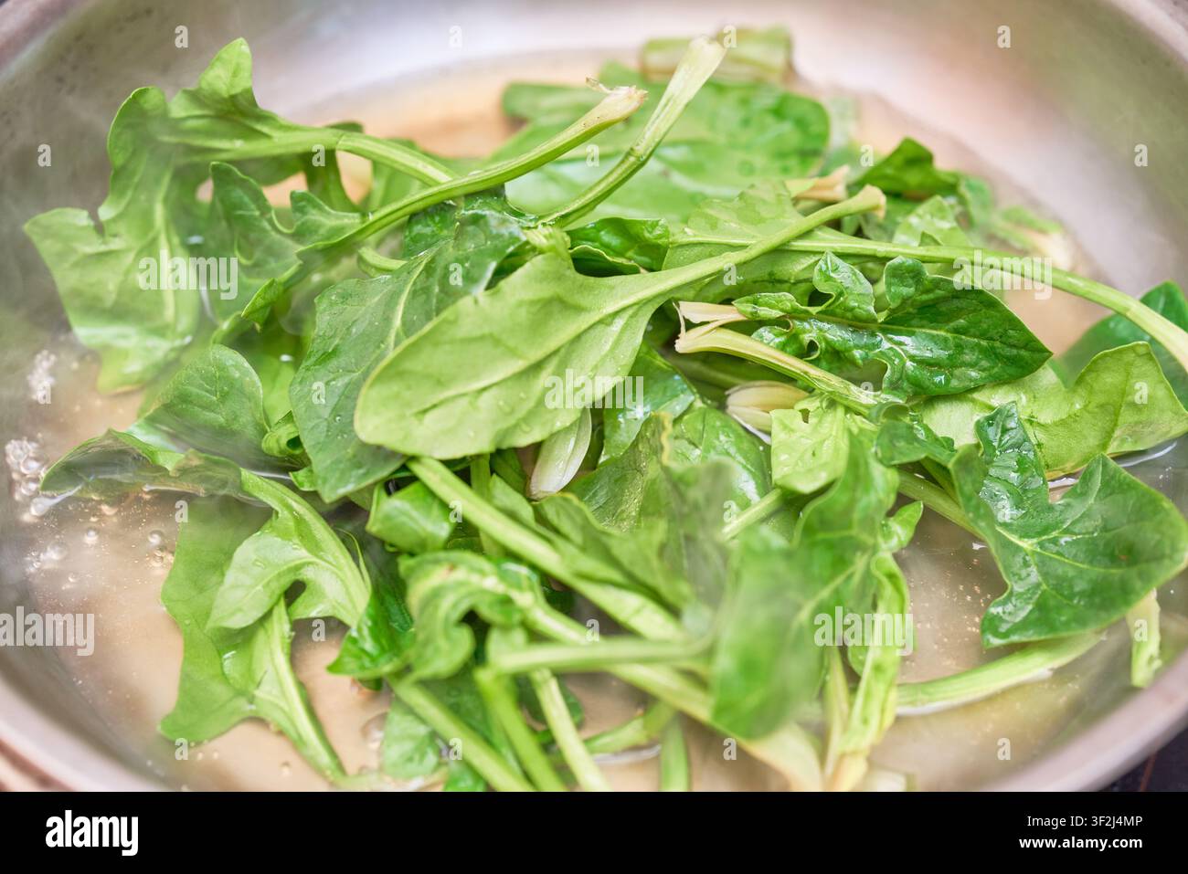 Foglie di spinaci fresche sbollentate in una padella in acciaio inossidabile che sobbollisce, perfette per guide di cucina sana e tutorial di ricette vegetariane. Foto Stock