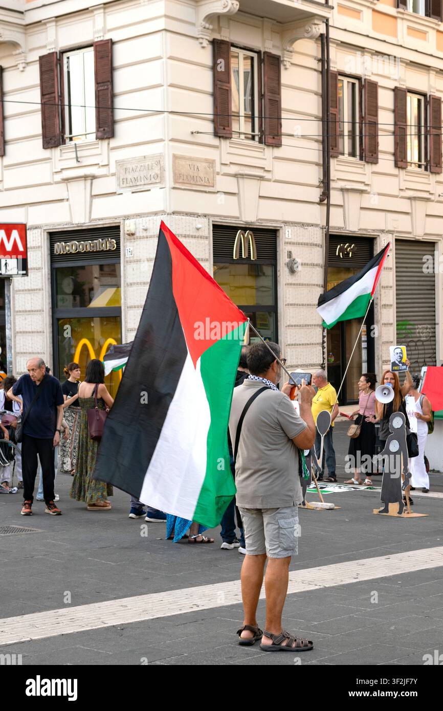 ROMA, ITALIA - 18 LUGLIO 2026: Un manifestante in piedi a un incrocio stradale, che solleva una grande bandiera palestinese durante una manifestazione di massa a Piazzale F Foto Stock