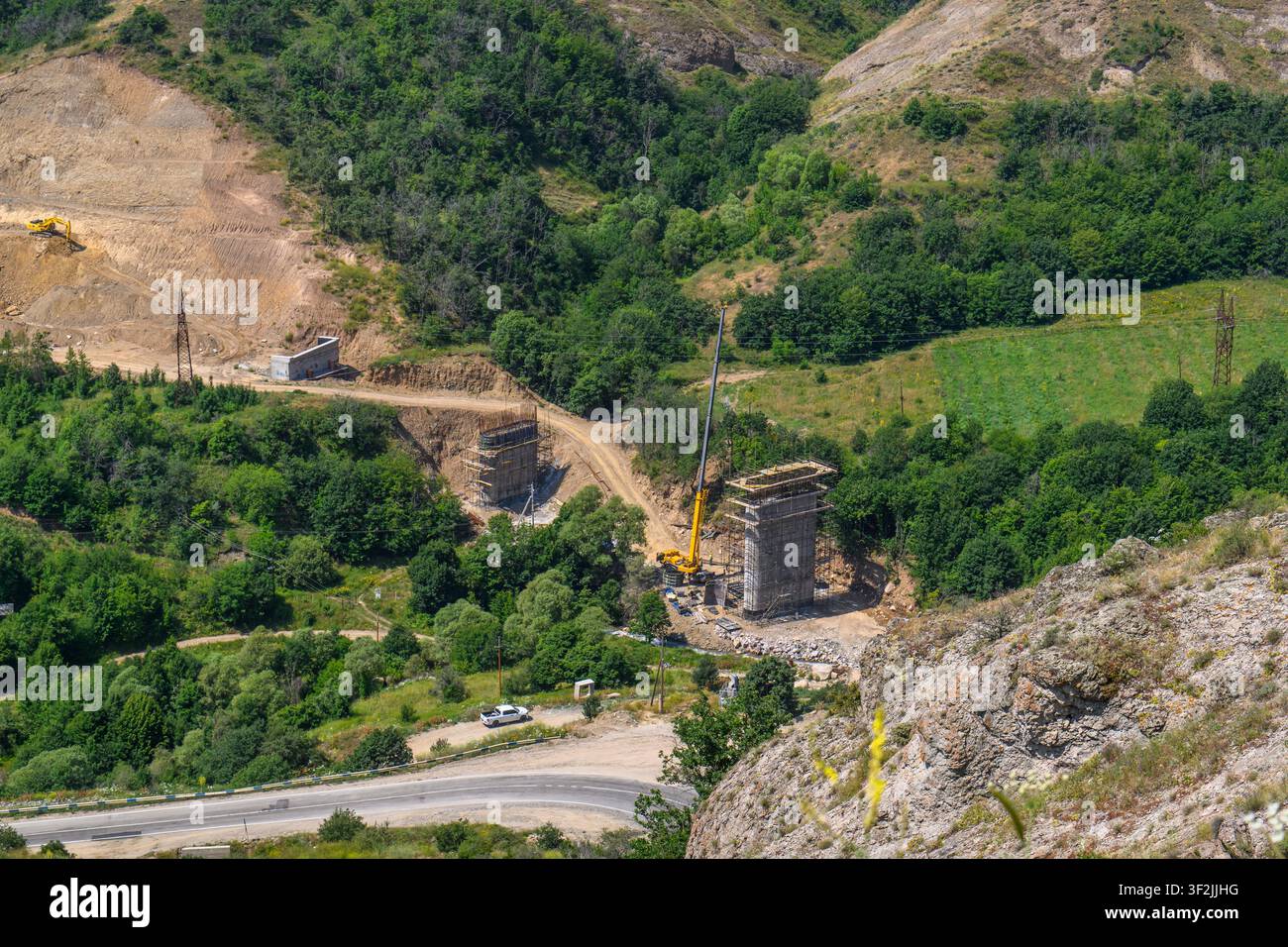 Grande gru per costruzioni solleva materiali vicino a vecchie rovine di pietra. Una foresta verde circonda la valle con una strada tortuosa. Sentieri sterrati collegano gli edifici Foto Stock