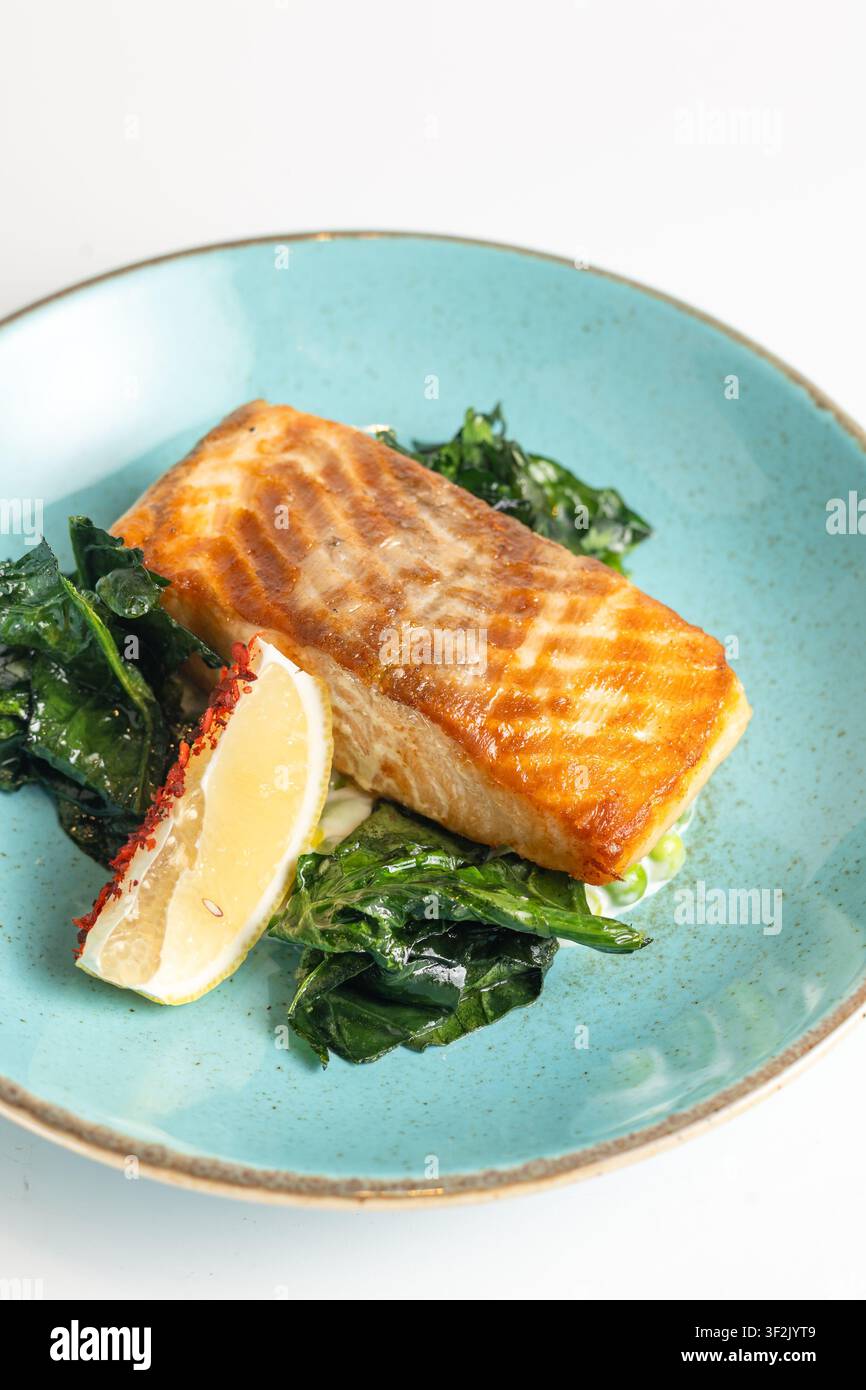 Filetto di salmone grigliato appetitoso con segni di griglia su un piatto di ceramica turchese con spinaci appassiti, fetta di limone e spezie rosse Foto Stock