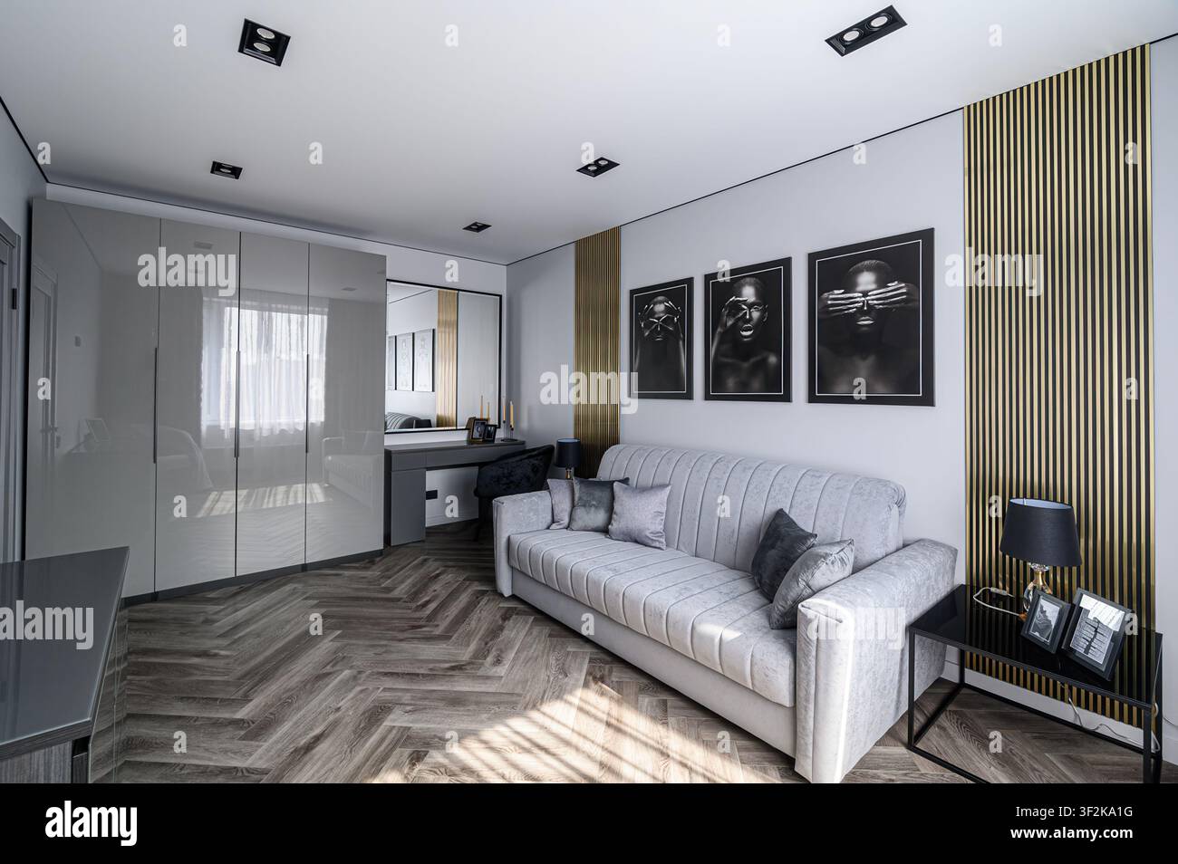 Stanza moderna con divano grigio, pavimenti in legno e arte monocromatica. Gli accenti dorati aggiungono eleganza. Gli armadi lucidi arricchiscono lo spazio Foto Stock