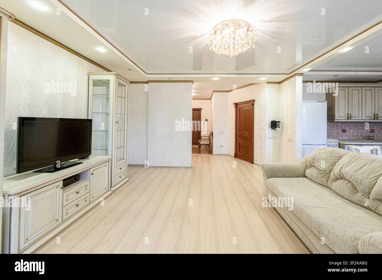 Soggiorno elegante con pavimenti in legno chiaro, una TV, un armadio, un divano e un lampadario di cristallo. Spazio caldo e accogliente Foto Stock