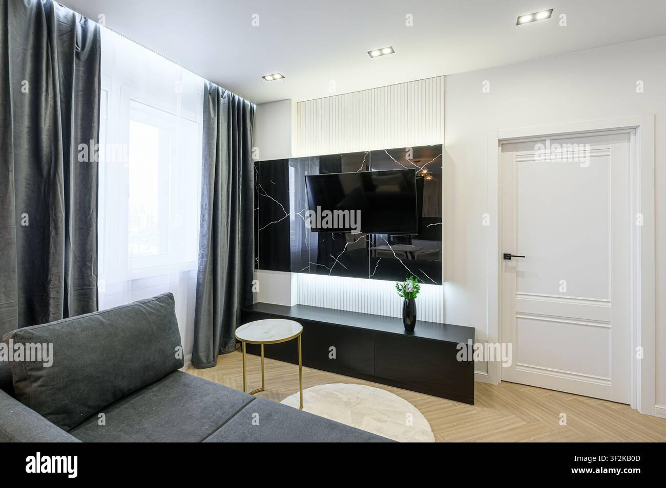 Interno di soggiorno elegante con un divano grigio, una parete TV nera elegante con dettagli in marmo e tende grigie eleganti. La stanza è ben illuminata e ha Foto Stock