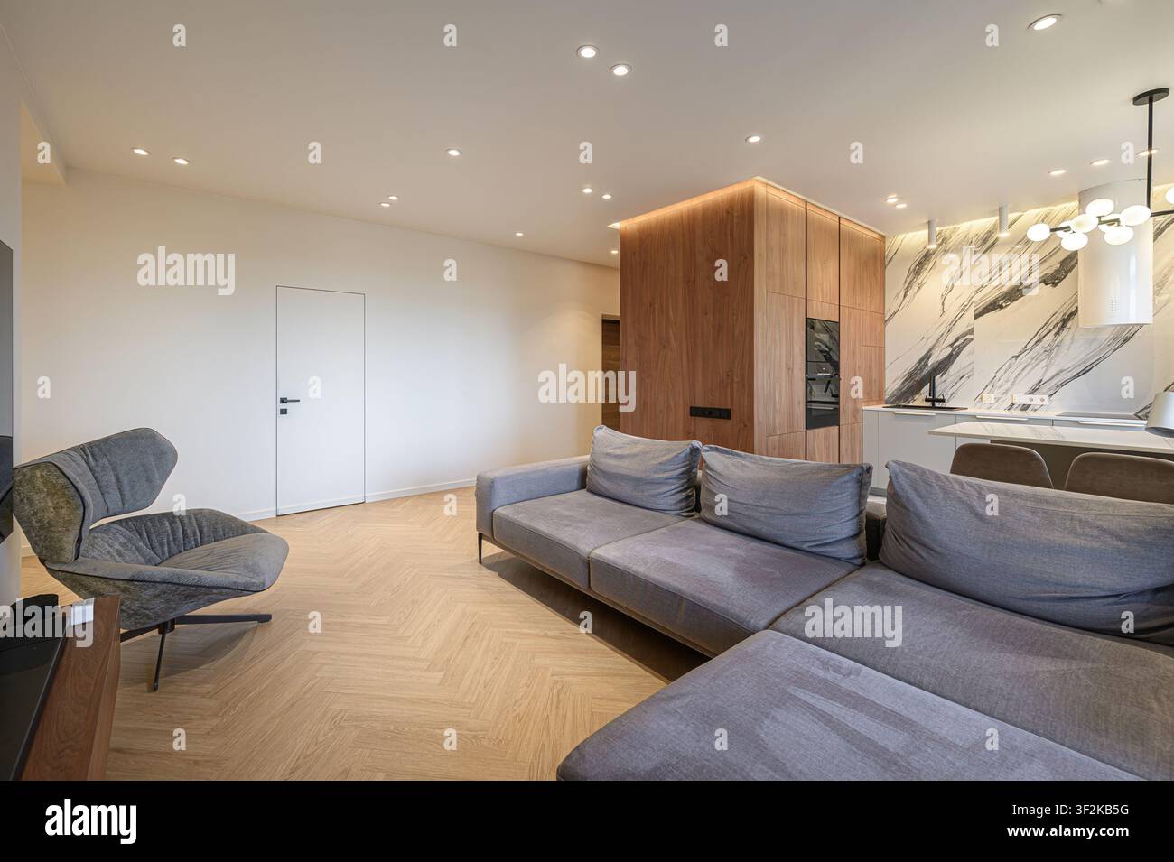 spazio abitativo moderno con divano ad angolo grigio, poltrona unica, pavimenti in legno e cucina contemporanea con dettagli in marmo Foto Stock