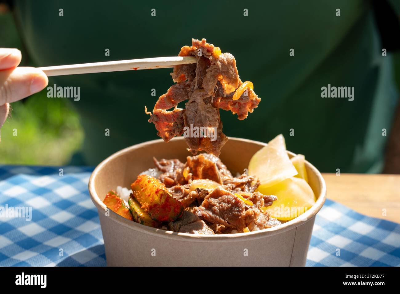 Bulgogi di manzo coreano in ciotola di carta. Riso, carne piccante e cetriolo marinato su uno sfondo a quadri in un caffè di strada in una giornata soleggiata, messa a fuoco selettiva, b Foto Stock