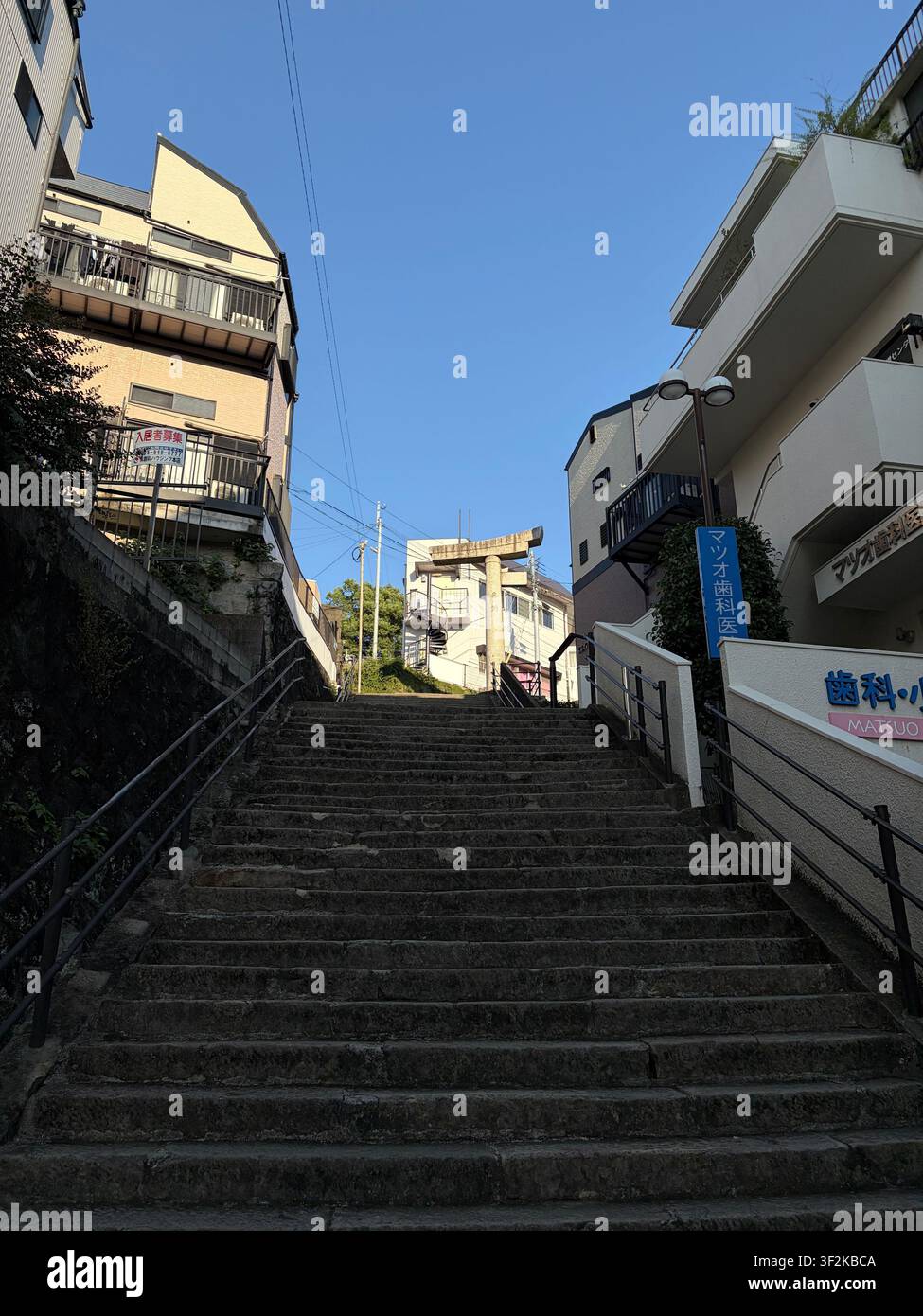 12 ottobre 2025, Nagasaki, Giappone – Scala di pietra ripida che sale attraverso un quartiere residenziale denso con insegne di negozi giapponesi e pali della luce sotto un cielo blu terso. - Immagine stock catturata con smartphone