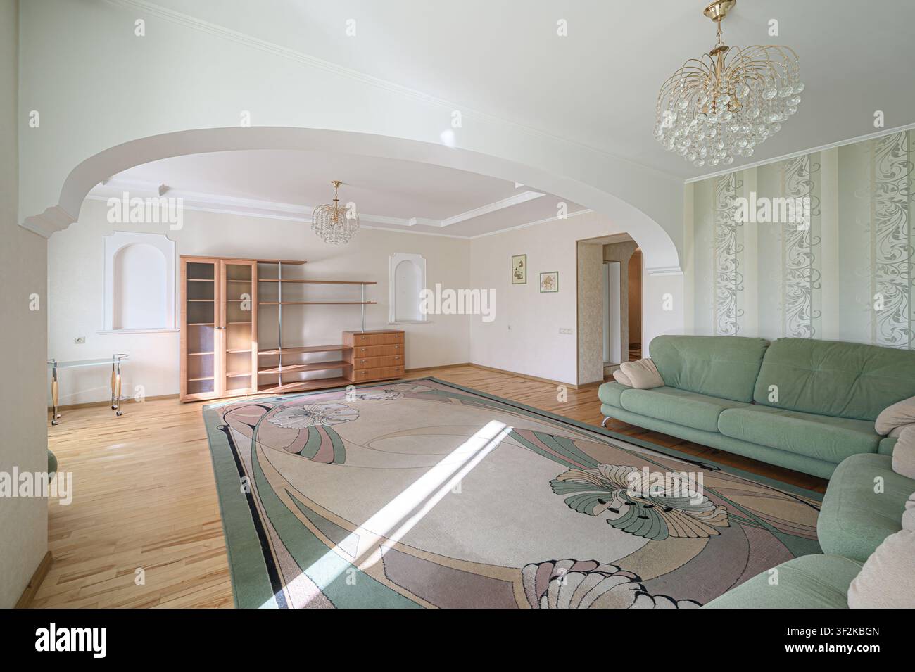 Soggiorno luminoso con un tappeto floreale, divano verde, arco e pavimenti in legno. Lampadari di cristallo forniscono luce Foto Stock