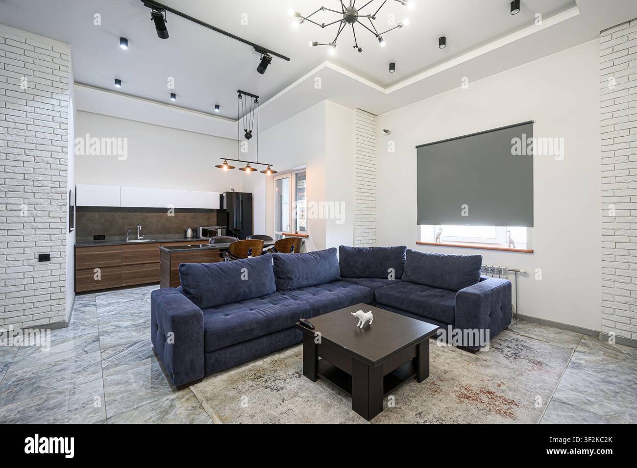 Spazio abitativo moderno con divano angolare blu, dettagli in legno scuro e area cucina aperta. Interno luminoso e stilizzato Foto Stock