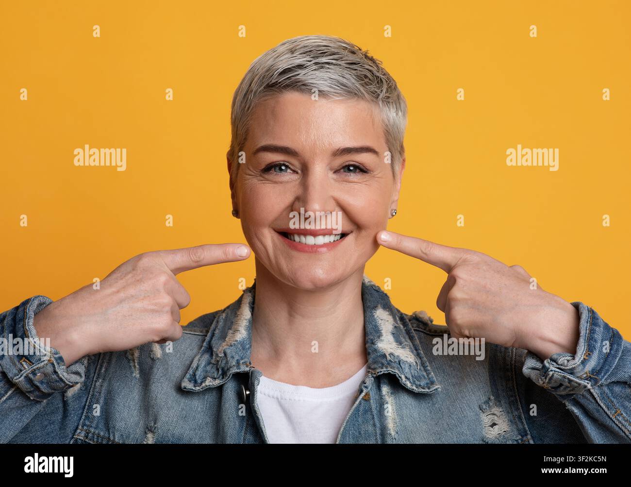 Smile perfetto. Donna matura che punta su denti sani forti, sfondo giallo Foto Stock