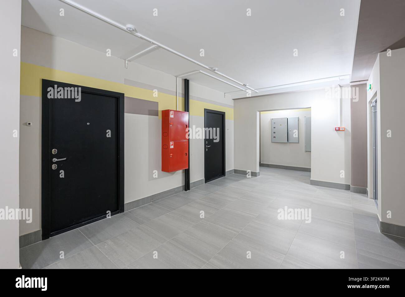Scatto interno di un corridoio di appartamento con porte nere, un armadietto antincendio rosso, pavimento piastrellato grigio e pareti nei toni neutri con accenti di colore Foto Stock