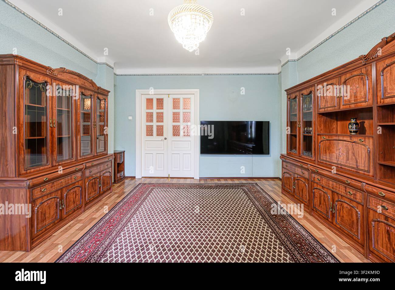 Una stanza ben illuminata presenta due grandi armadi in legno, una TV montata sopra un tappeto e un lampadario di cristallo. Una porta bianca è centrata tra gli armadi Foto Stock
