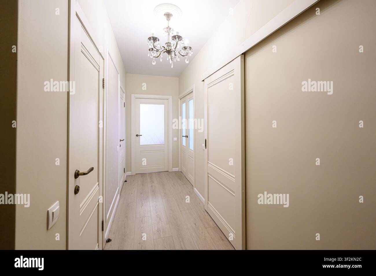 corridoio ben illuminato con porte bianche e pavimenti in legno chiaro, illuminato da un lampadario decorativo Foto Stock