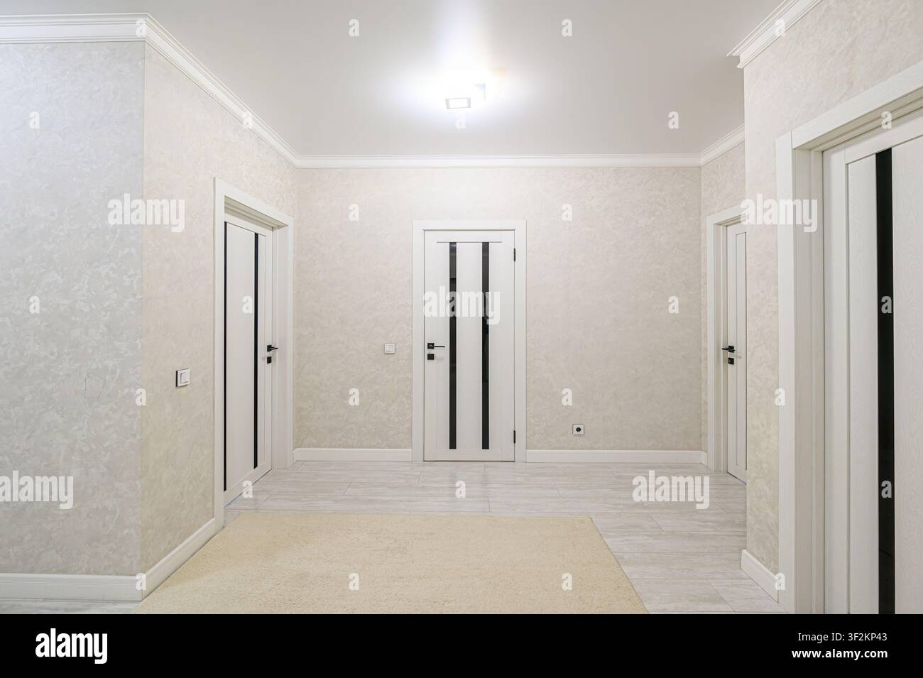 corridoio luminoso con tre porte bianche con dettagli neri, pareti a texture leggera, un tappeto neutro e un pavimento piastrellato, illuminato da un lampadario centrale Foto Stock