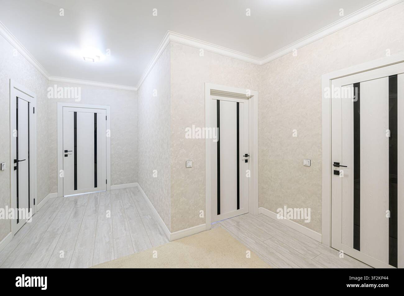 corridoio luminoso con tre porte bianche con dettagli neri, pareti a texture leggera, un tappeto neutro e un pavimento piastrellato, illuminato da un lampadario centrale Foto Stock