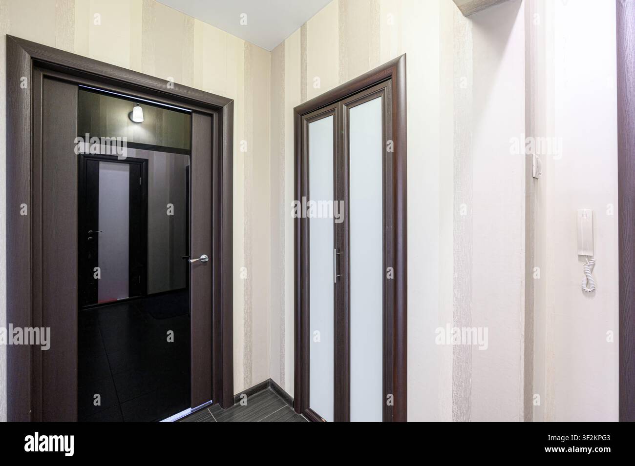 Foto che mostra un corridoio con porte. Una porta è aperta, rivelando una stanza buia. Il corridoio ha carta da parati a strisce e un pavimento piastrellato grigio scuro Foto Stock