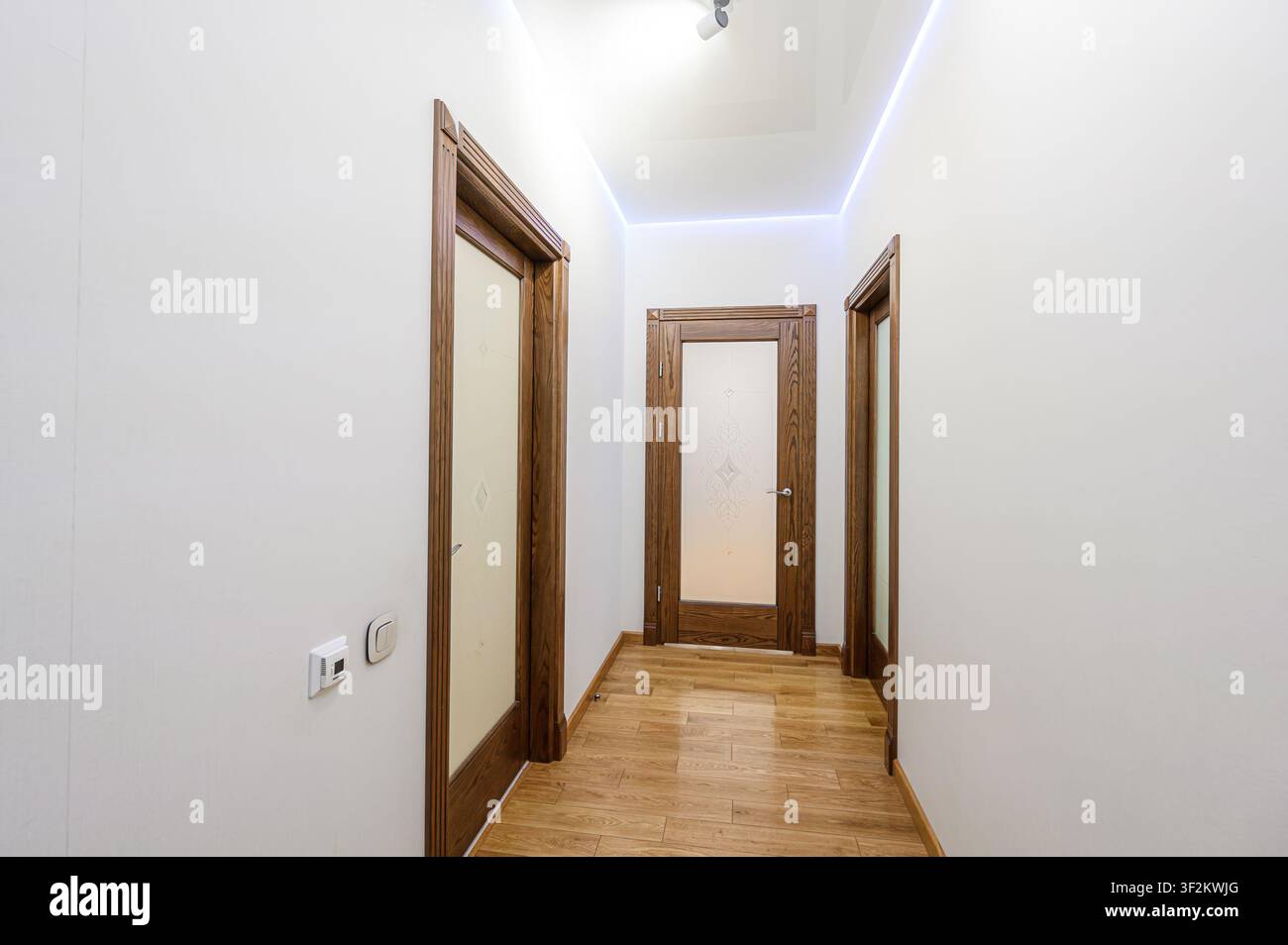 Corridoio con porte di legno e pannelli di vetro, pareti bianche e pavimenti in legno. Il soffitto ha luci incassate e una striscia di illuminazione a LED Foto Stock