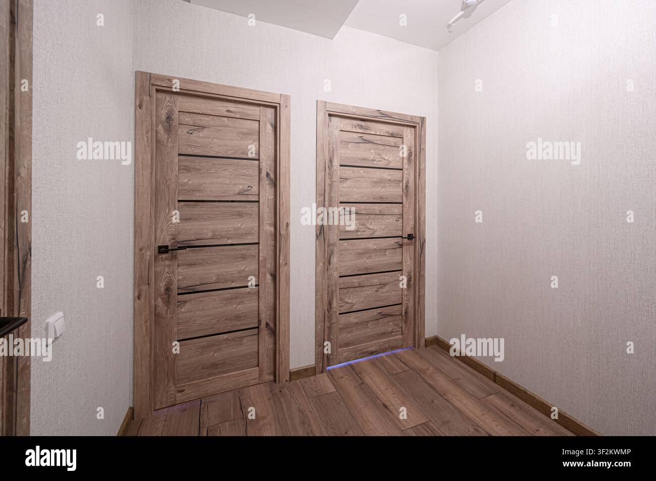 fotografia interna ben illuminata che mostra due porte moderne in legno con accenti orizzontali neri su pareti bianche ruvide. Il pavimento in legno si estende nella Foto Stock