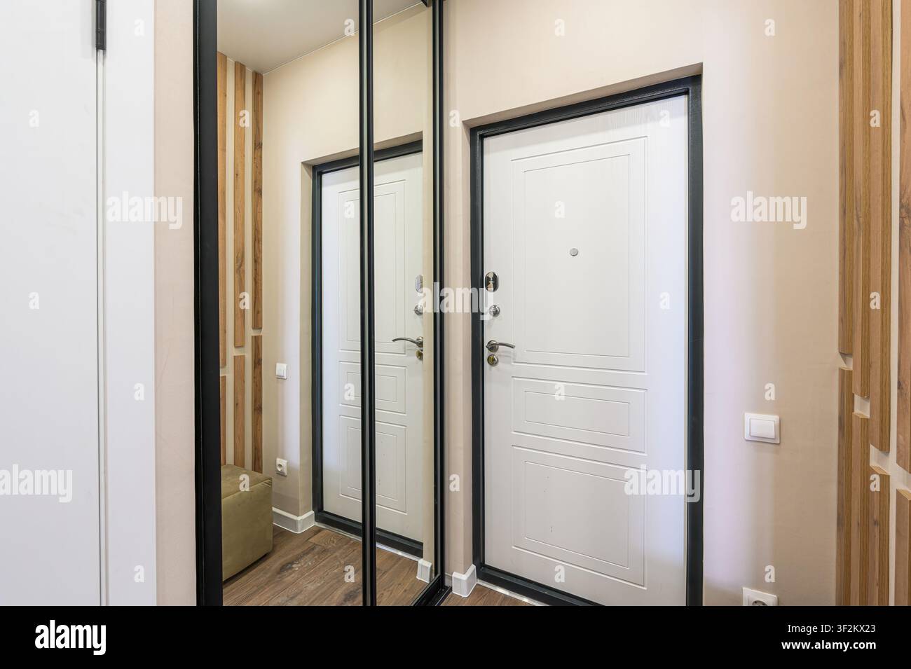 Un colpo d'interno mostra un ingresso moderno con una porta bianca incorniciata in nero, ante di armadio a specchio, dettagli in legno e pareti beige chiaro Foto Stock