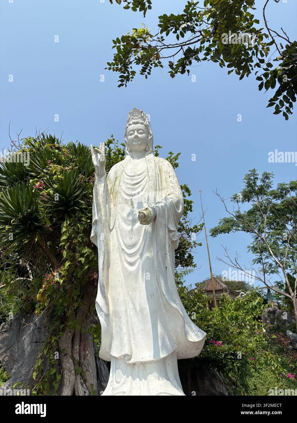 7 aprile 2025, Da Nang, Vietnam - Statua di bodhisattva Guanyin (Quan Am) in marmo bianco che si erge sopra la vegetazione tropicale contro un cielo blu terso. - Immagine stock catturata con smartphone
