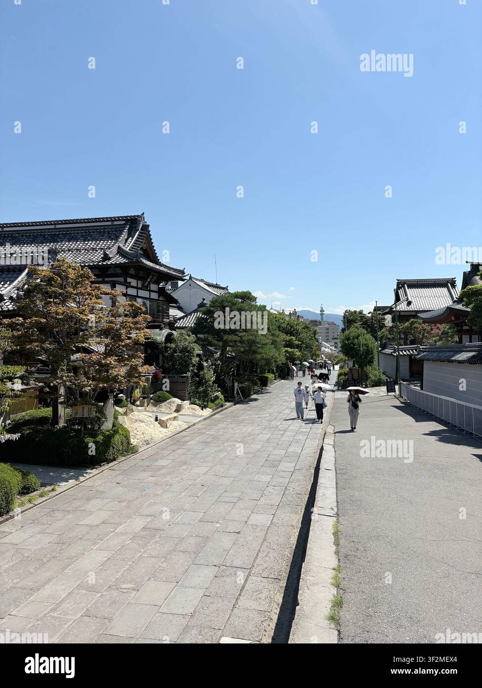 28 agosto 2025, Nagano, Giappone – Muro di pietra lungo un complesso templare tradizionale giapponese con tetti di pagoda e alberi da giardino, visitatori a passeggio. - Immagine stock catturata con smartphone
