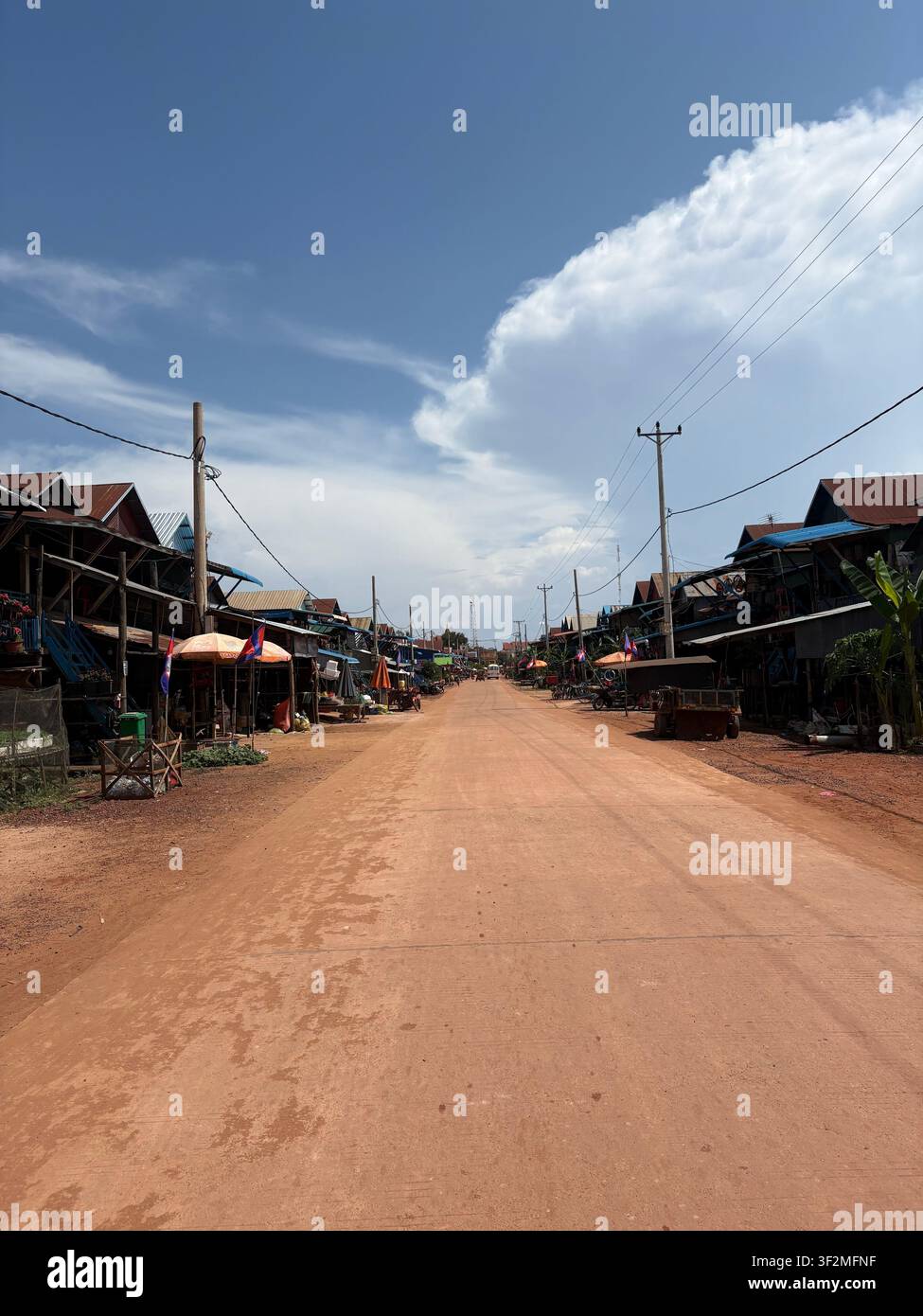 18 aprile 2025, Prasat Bakong, Siem Reap, Cambogia – Strada di un villaggio rurale con strada di terra rossa, case di legno su palafitte, linee elettriche e bandiere cambogiane sotto un cielo blu. - Immagine stock catturata con smartphone